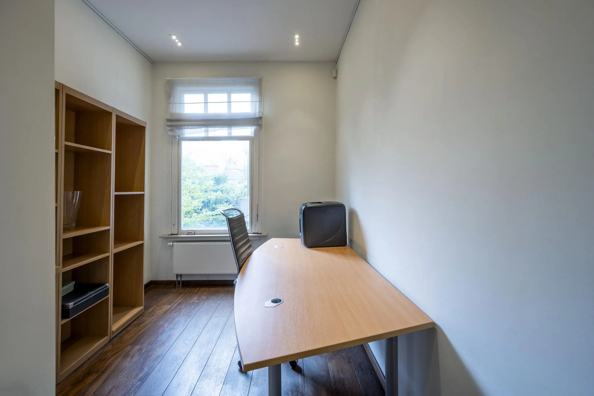 Kantoorruimte aan de Lomanstraat met houten vloer, bureau, bureaustoel, open kast en raam met uitzicht op groen.