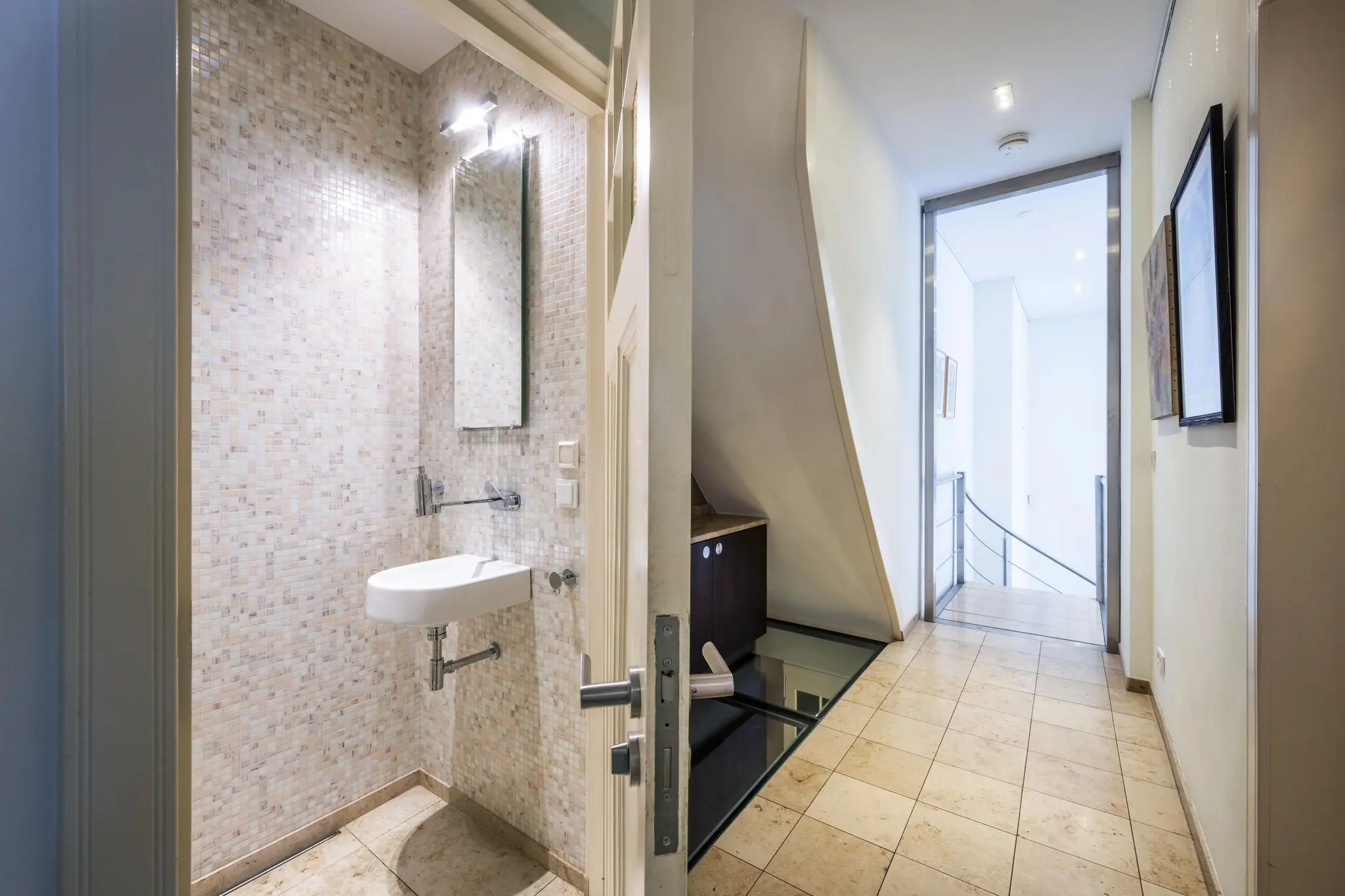 Moderne gang met een open deur naar een kleine toiletruimte met mozaïektegels en een zwevend fonteintje.