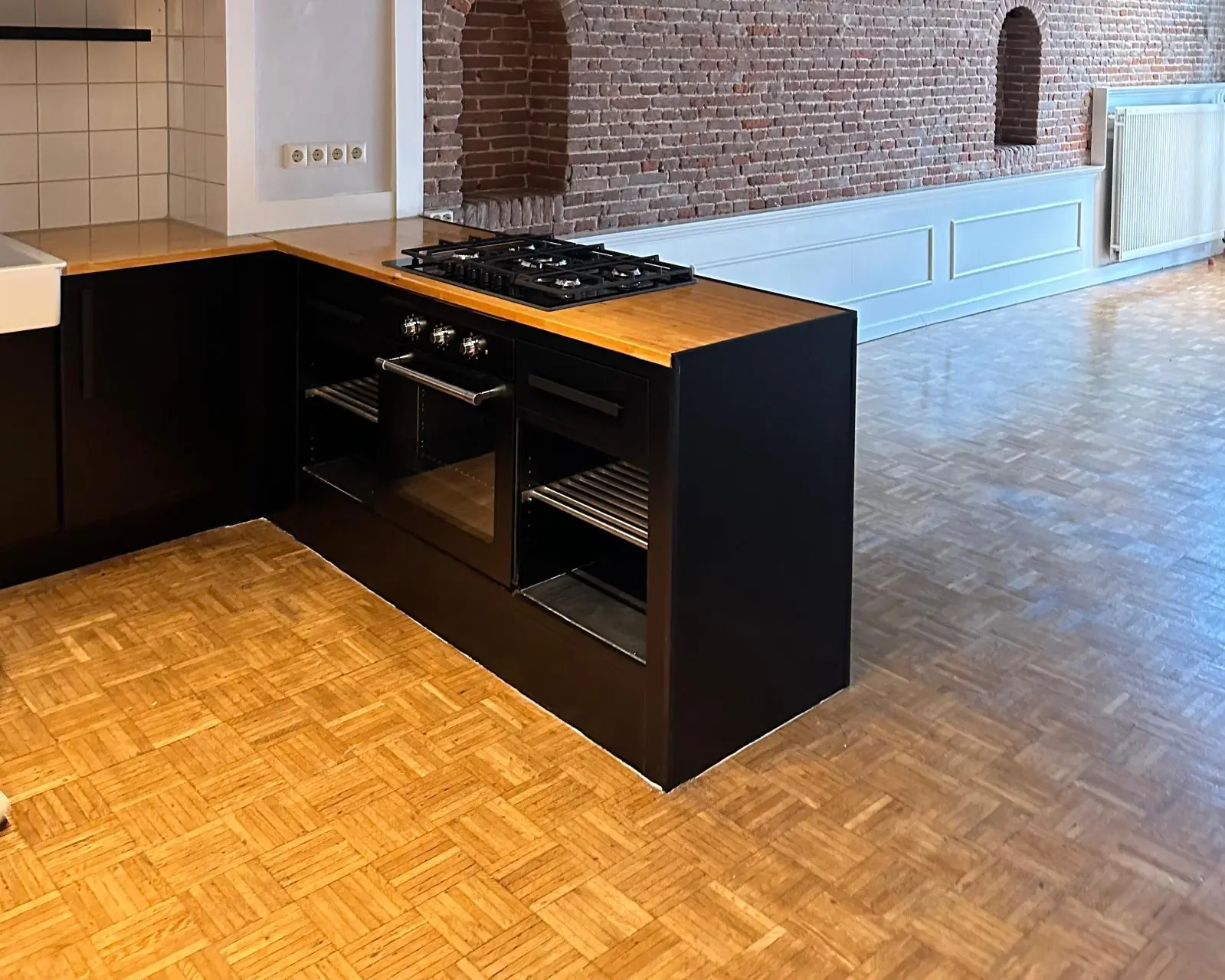 Moderne zwarte hoekkeuken met houten aanrechtblad in een appartement aan de Herengracht, met zicht op een bakstenen muur en visgraatparketvloer.