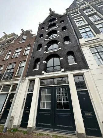 Gevelaanzicht van een smal, monumentaal pand aan de Herengracht in Amsterdam met zwarte bakstenen en witte kozijnen.