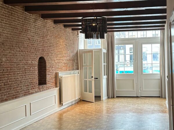 Lichte woonkamer aan de Herengracht met een bakstenen muur, houten balkenplafond en grote ramen met uitzicht op de gracht.