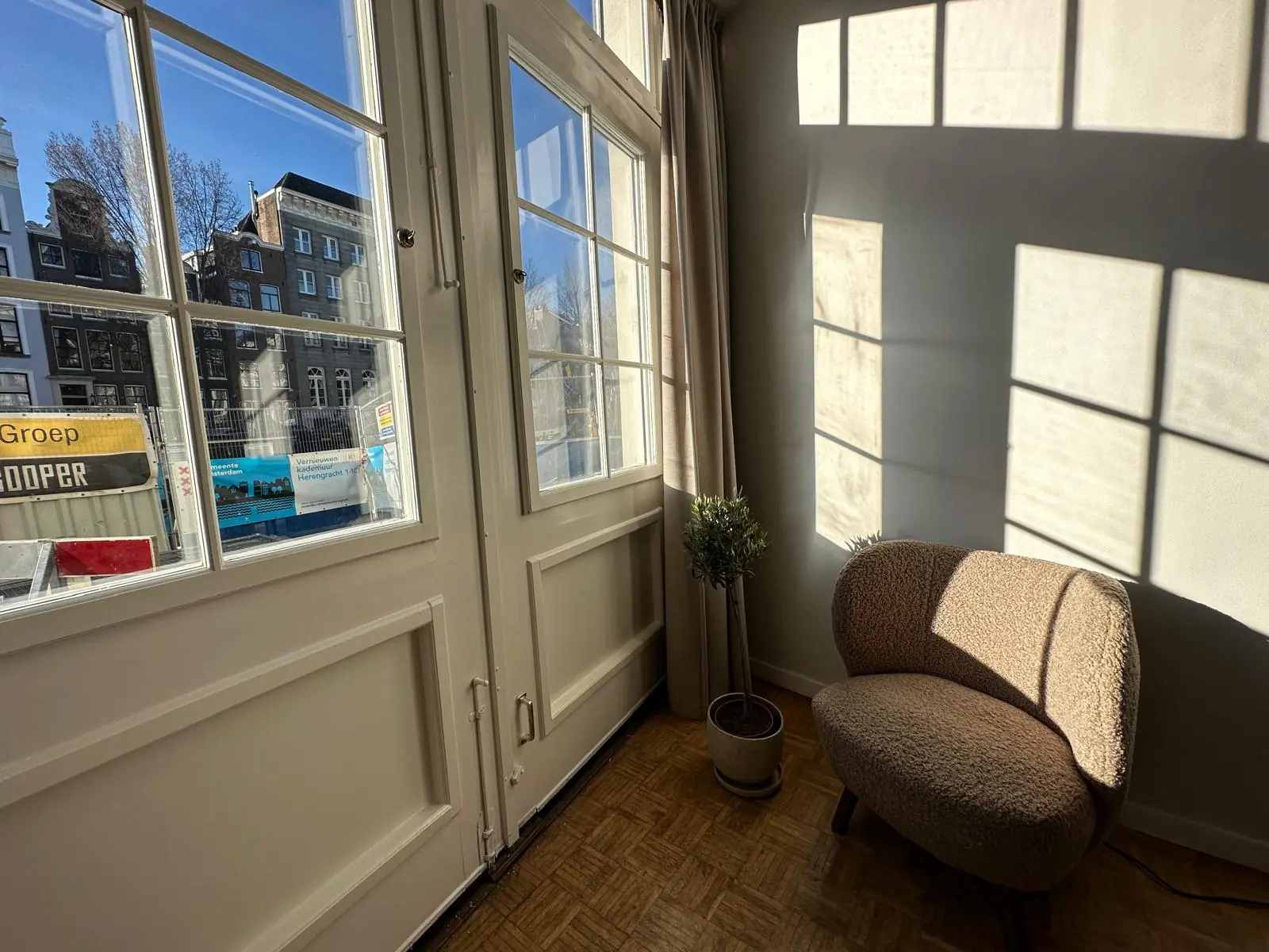 Zonnige zithoek bij een raam met uitzicht op de Herengracht en bouwactiviteiten buiten.