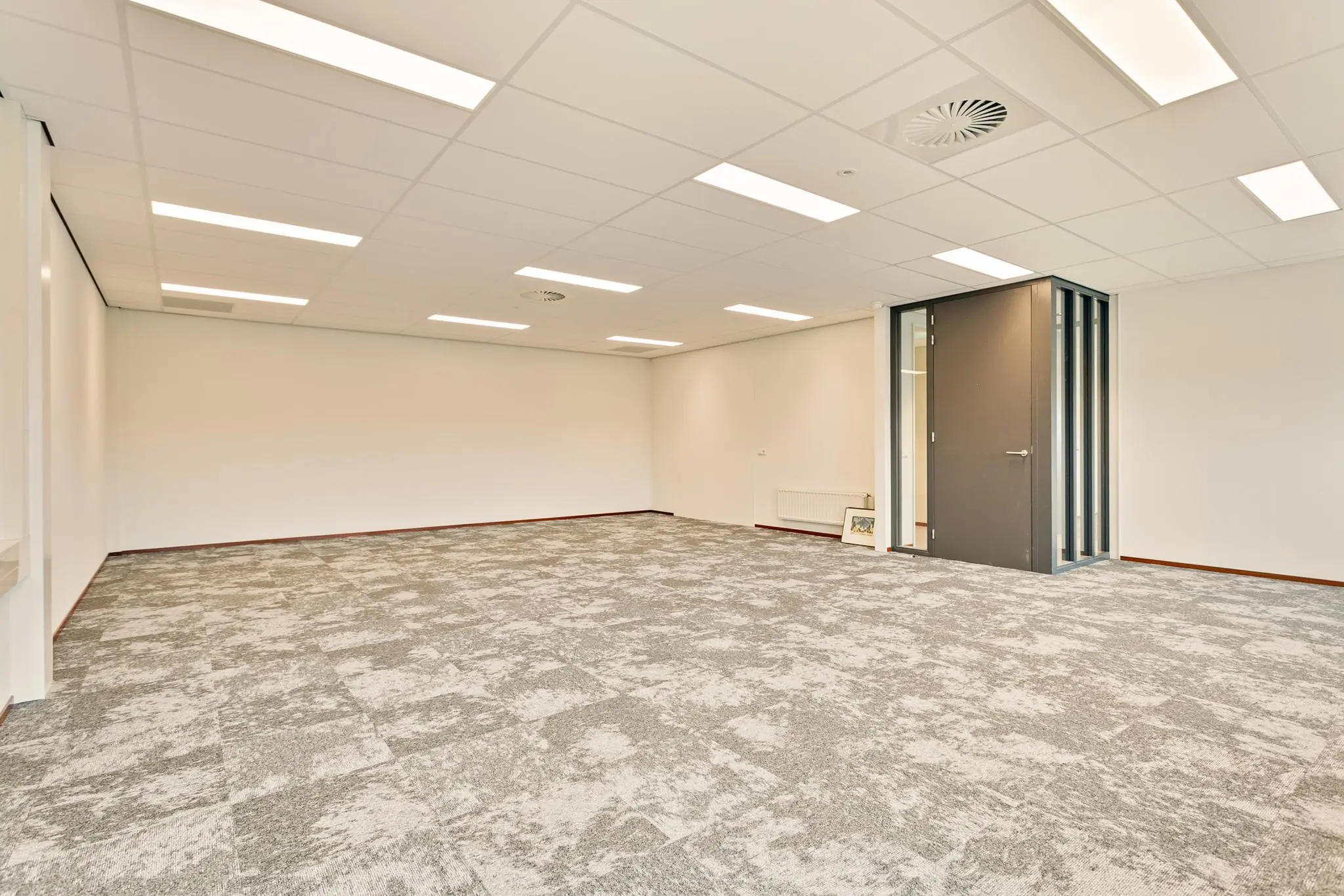 Lege kantoorruimte met grijs tapijt, witte wanden en een gesloten donkere deur.