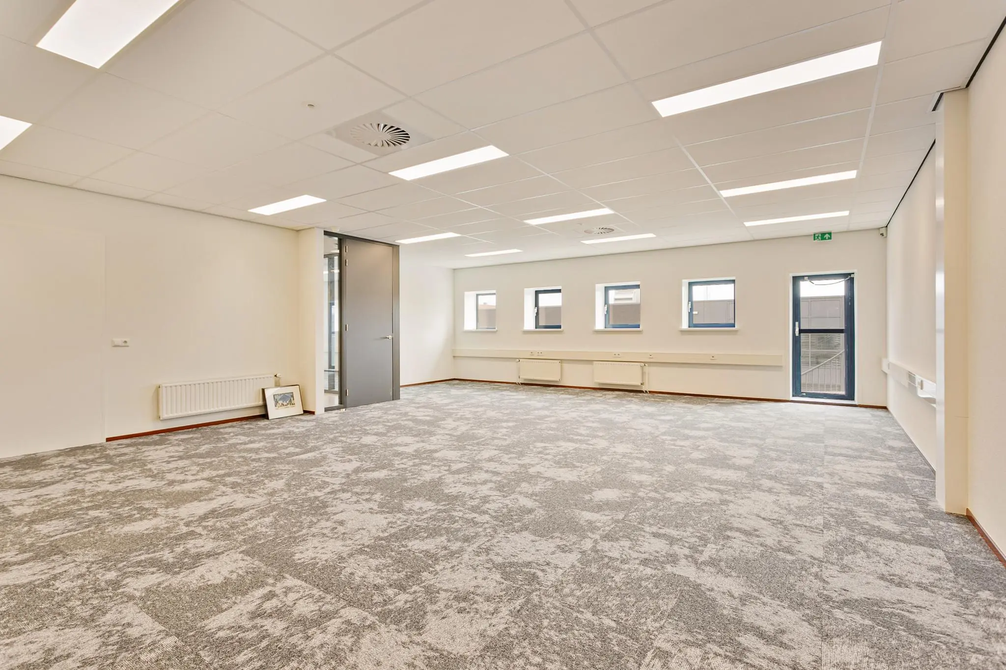 Lichte, lege kantoorruimte met grijs tapijt, witte muren en meerdere ramen.