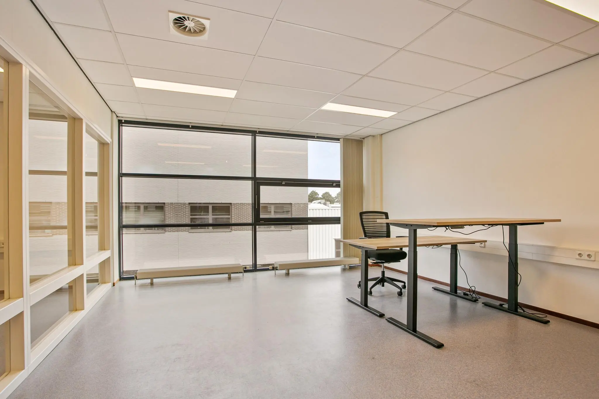 Licht en leeg kantoor met een verstelbaar bureau, bureaustoel en grote ramen met uitzicht op een andere gebouwvleugel.