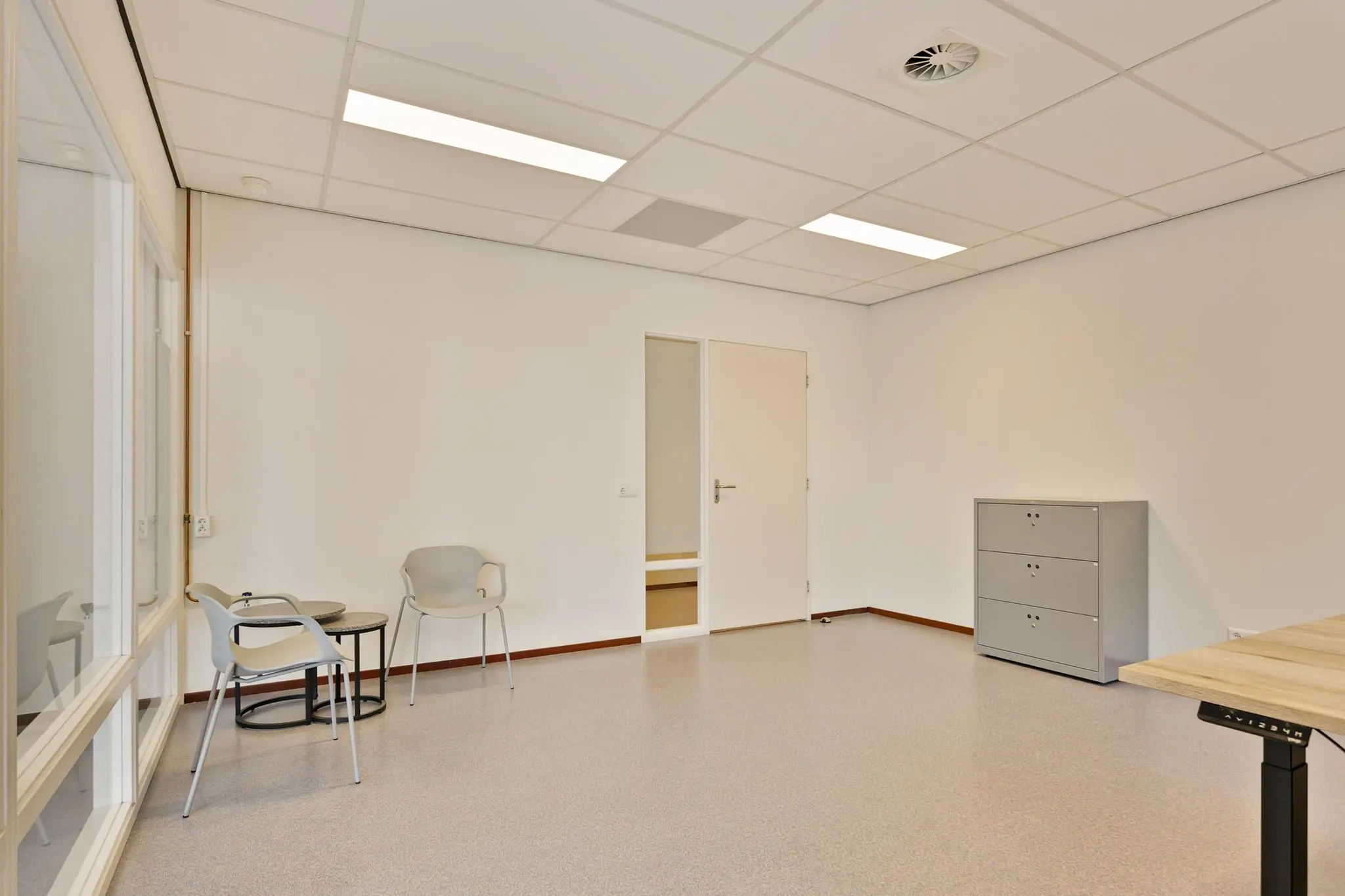 Een lege, moderne kantoorruimte met twee stoelen, een klein tafeltje, een ladekast en een deels geopend bureau.