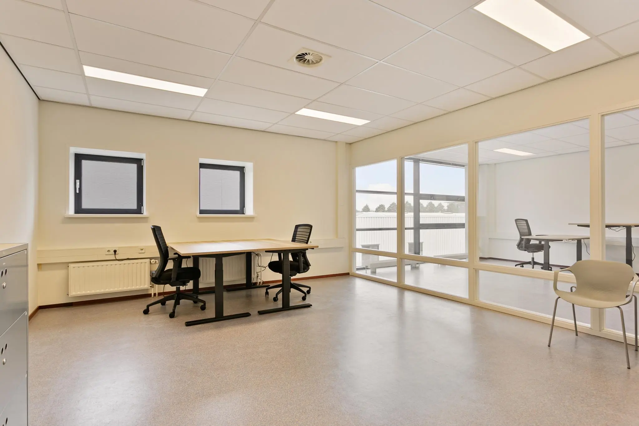 Lichte kantoorruimte met bureaus, bureaustoelen en een glazen tussenwand naar een aangrenzende ruimte.