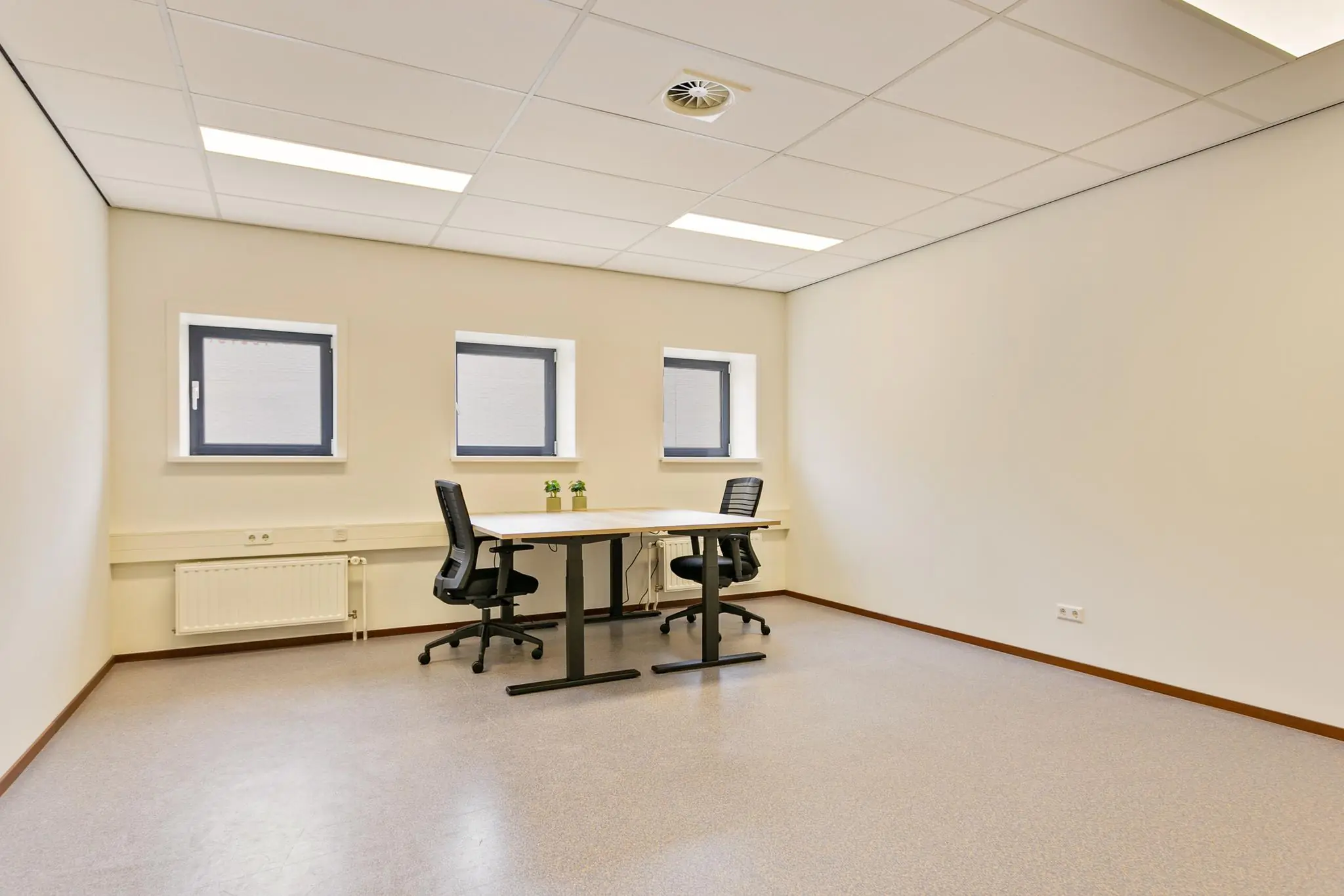 Twee bureaustoelen staan tegenover elkaar aan een eenvoudig bureau in een licht en minimalistisch kantoor met drie kleine ramen.