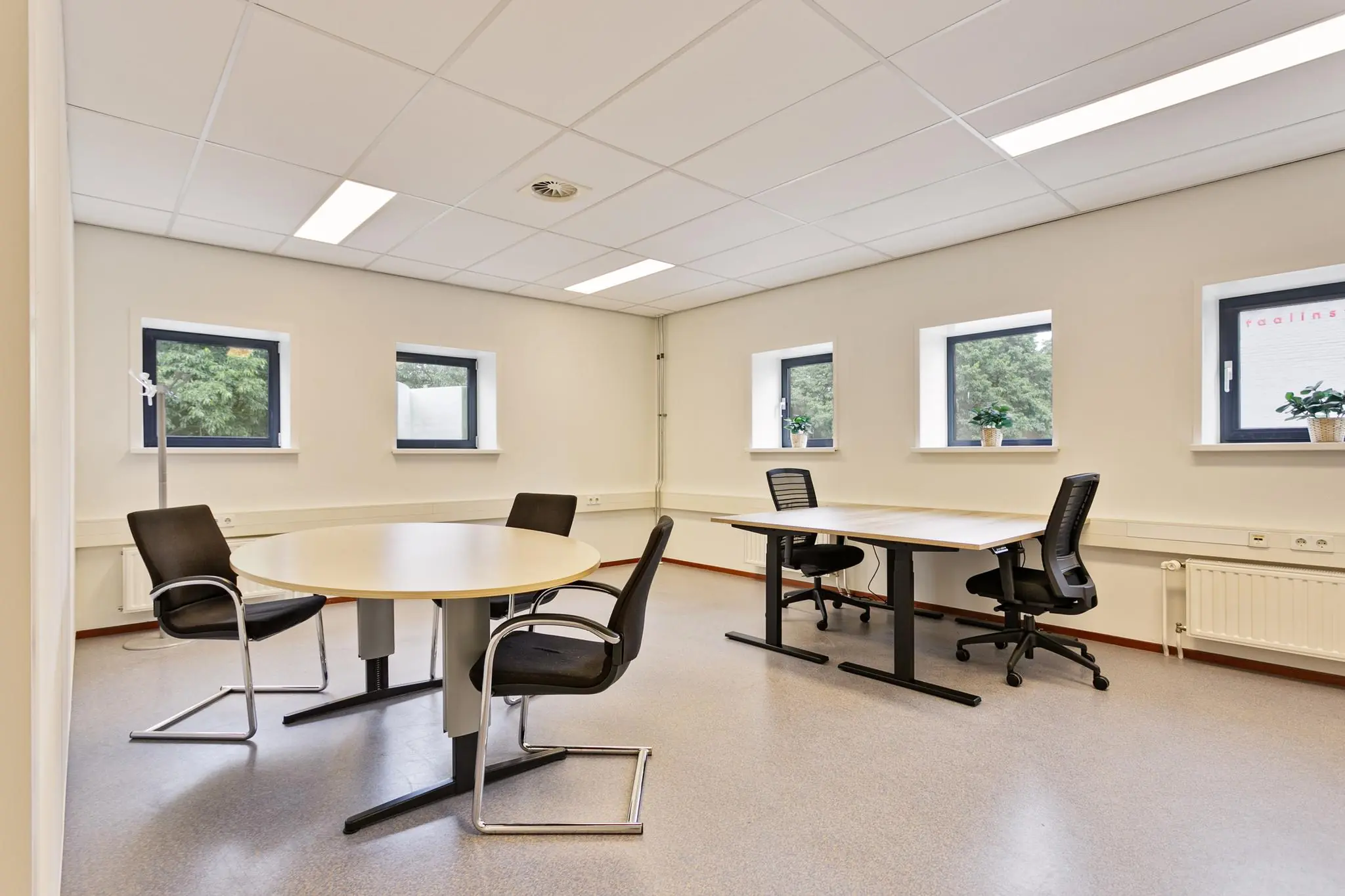 Licht en modern kantoor met ronde vergadertafel, twee bureaus en vier ramen met uitzicht op groen.