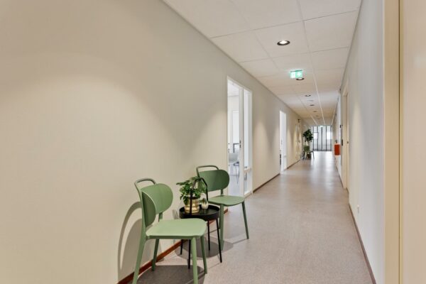 Lichte kantoorhal met twee groene stoelen en een tafeltje met planten tegen de muur.