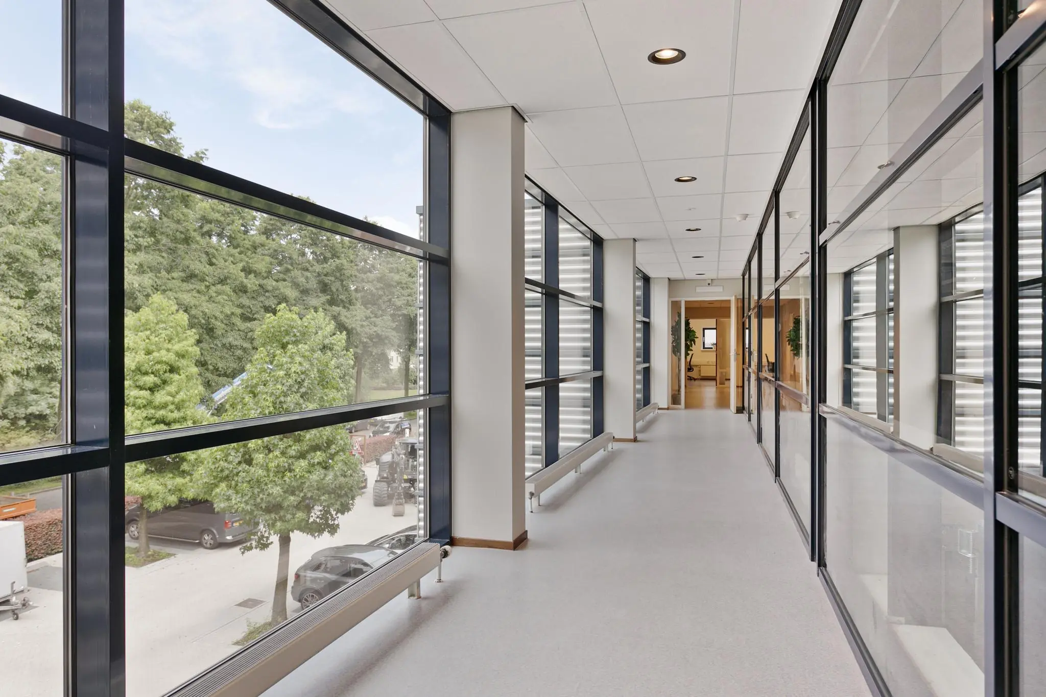Lichte kantoorgang met grote ramen en zicht op bomen en geparkeerde auto's buiten.