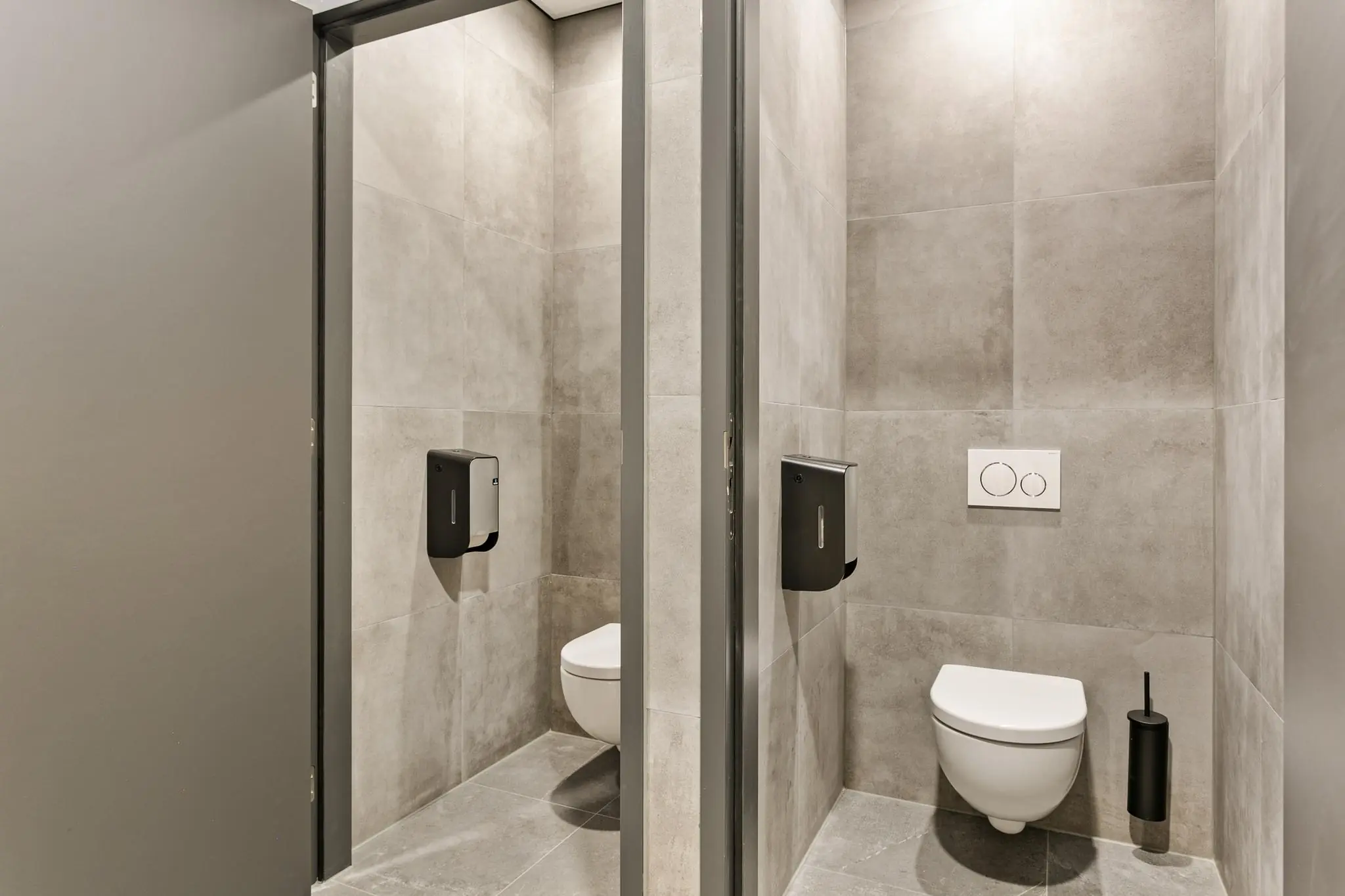 Twee moderne toiletcabines met grijze tegels, hangtoiletten en zwarte toiletrolhouders.