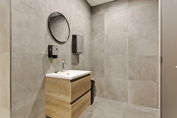 Moderne, grijze toiletruimte met houten wastafel, ronde spiegel en wanddispensers.