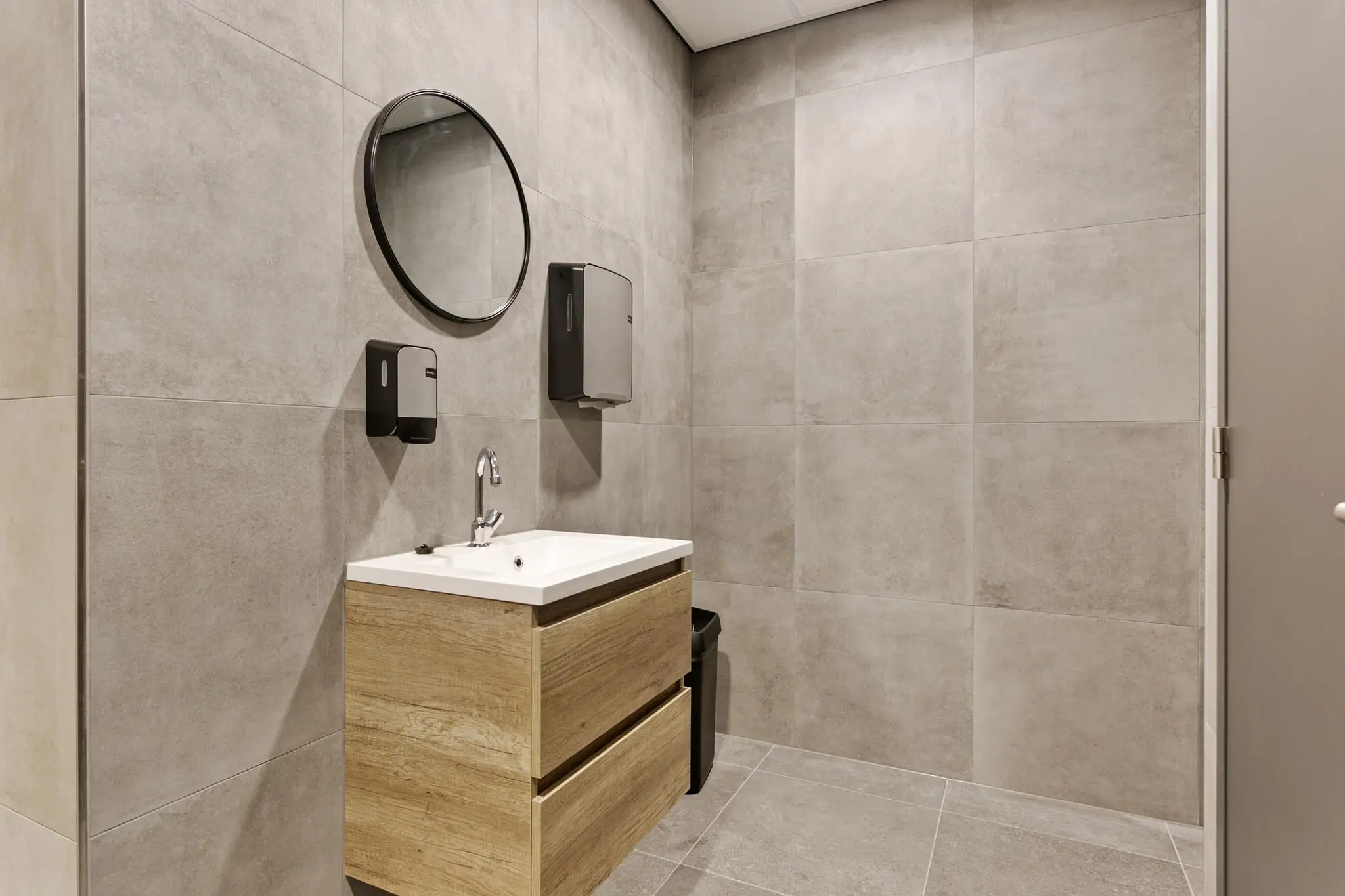 Moderne, grijze toiletruimte met houten wastafel, ronde spiegel en wanddispensers.