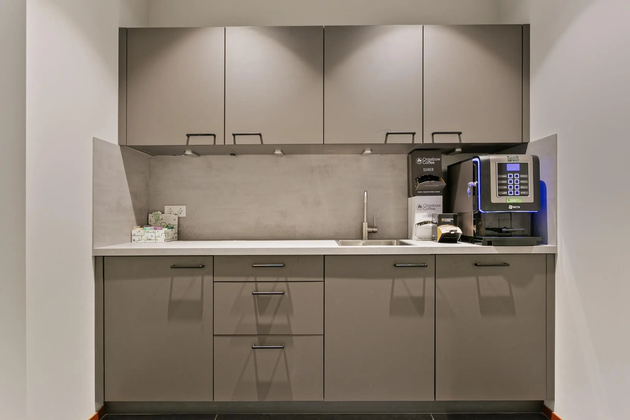 Moderne kantoorkeuken met koffieautomaat, wastafel en grijze keukenkastjes.