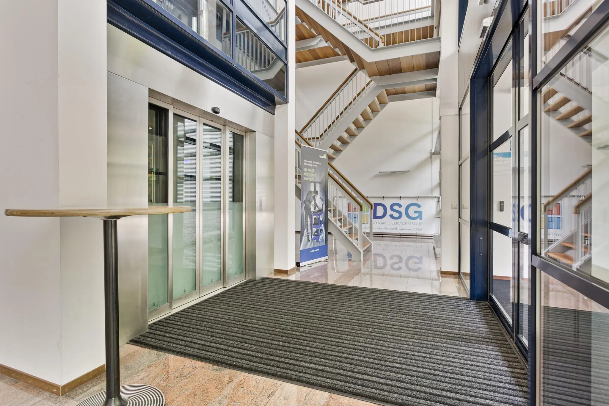 Moderne entreehal van een kantoorgebouw met glazen liftdeuren, trap en een banner van Dynamic Service Group (DSG).