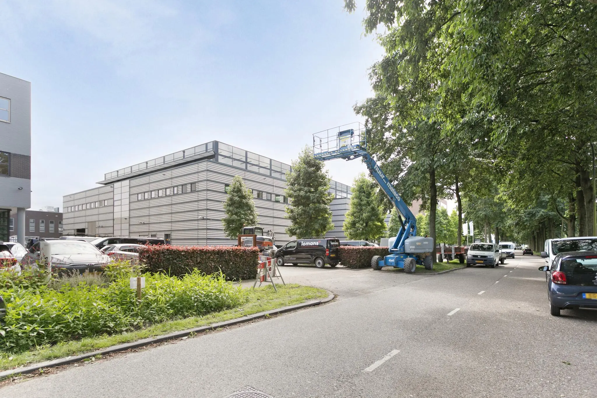 Een hoogwerker en bedrijfswagens staan geparkeerd naast een modern industriepand in een groene bedrijfsomgeving.
