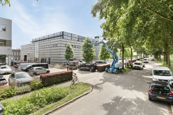 Moderne kantoorgebouwen met geparkeerde auto’s en een hoogwerker aan een rustige, met bomen omzoomde straat.