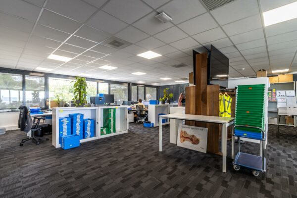 Kantoorruimte aan de Laanakkerweg met bureaus, kasten vol blauwe en groene opbergmappen en enkele werkende personen.