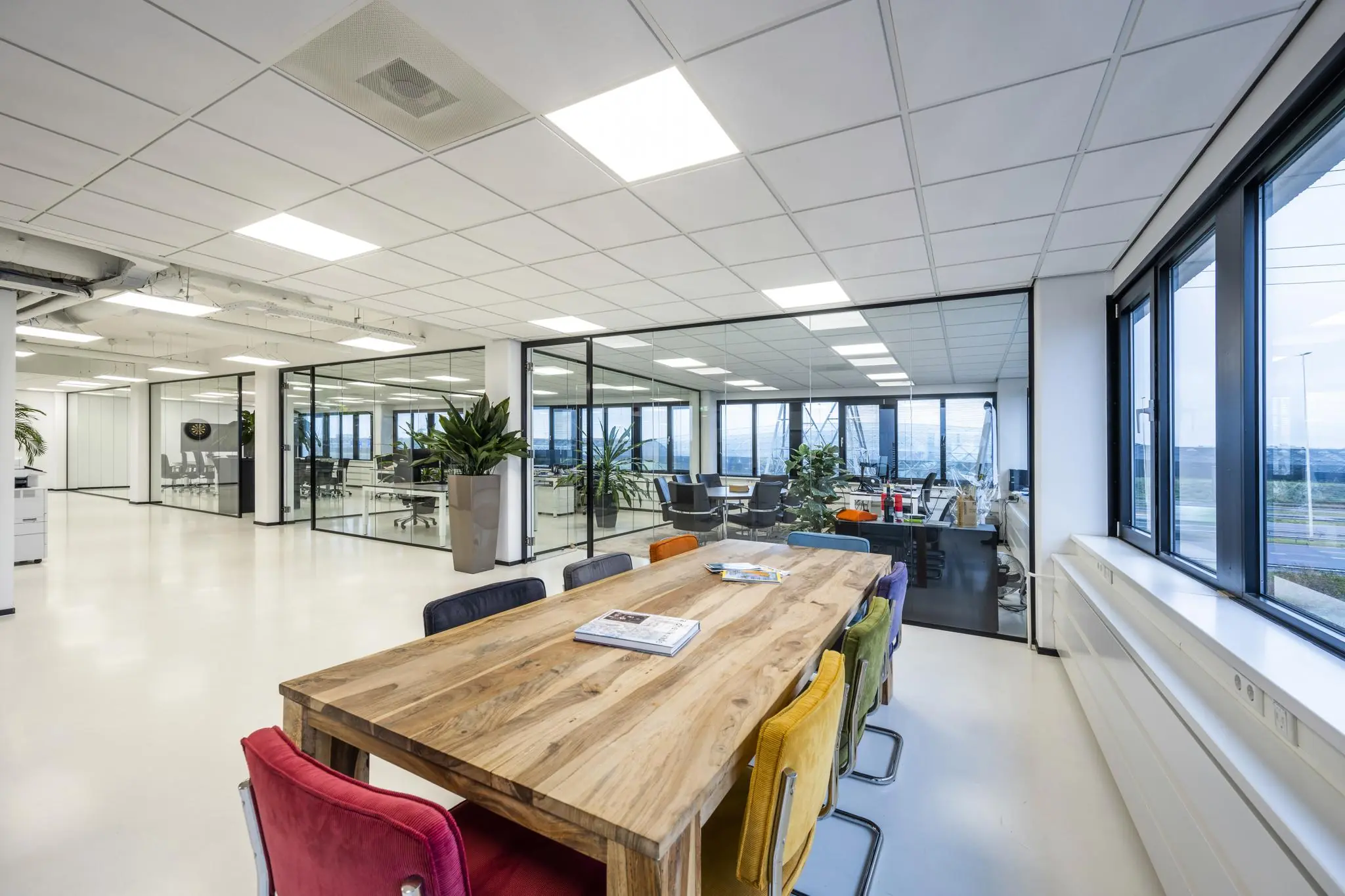 Moderne kantooruimte aan de Savannahweg met glazen vergaderruimtes en een houten tafel met kleurrijke stoelen.