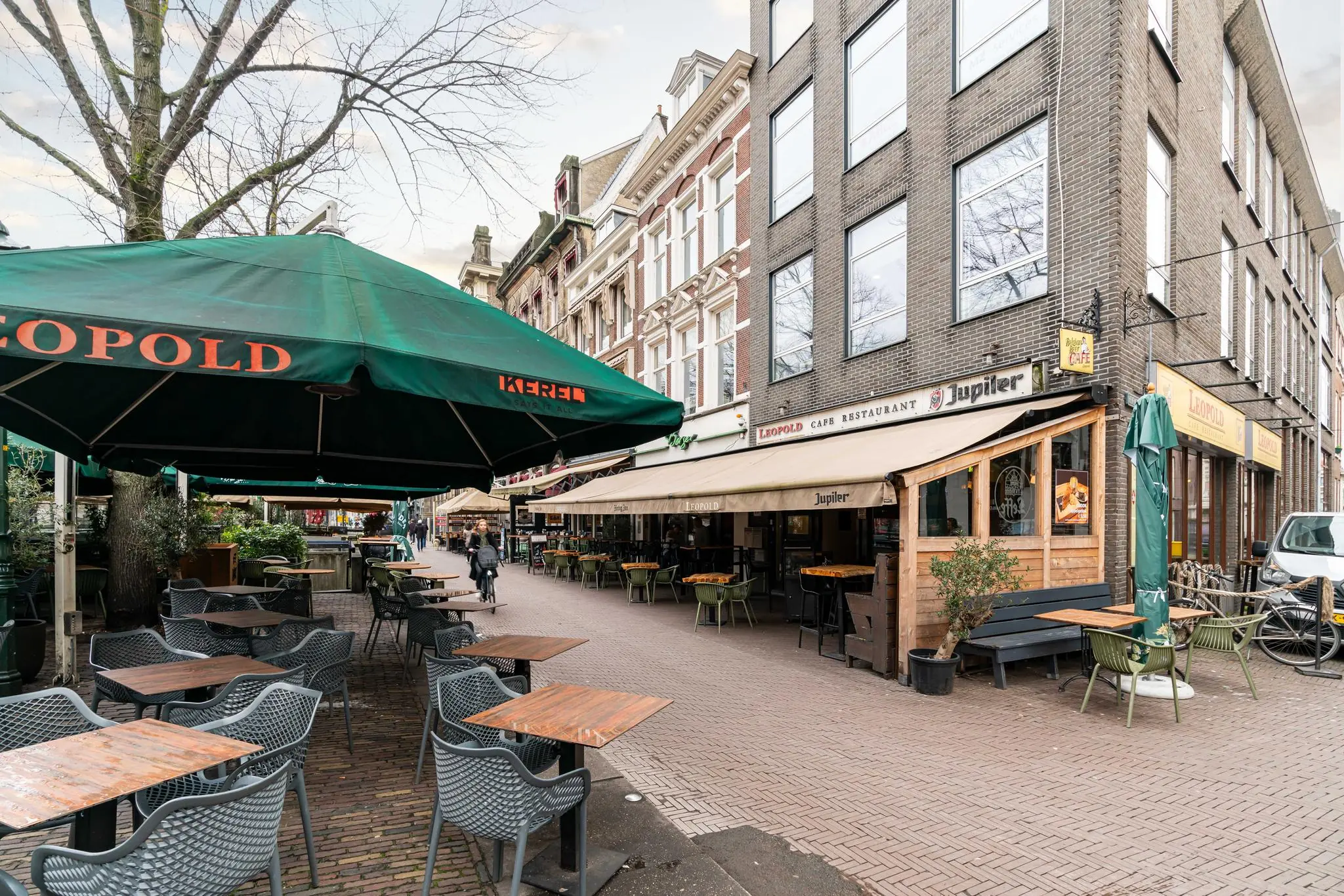 Terras van Café Restaurant Leopold aan een gezellig plein met tafels, stoelen en groene parasols.