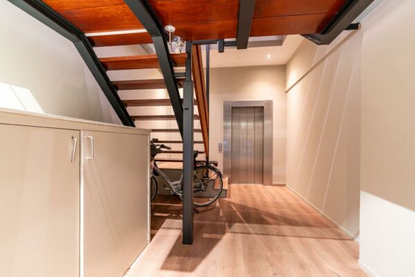 Moderne hal met houten trap, metalen liftdeur, een fiets en een opbergkast.