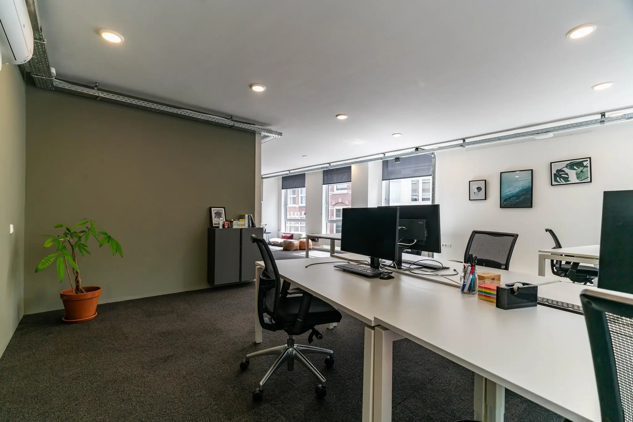 Modern kantoor met bureaus, beeldschermen, planten en wanddecoratie aan een grote, lichte raamzijde.