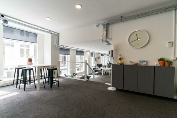 Moderne, open kantoorruimte met hoge ramen, een zithoek, planten en een wandklok.