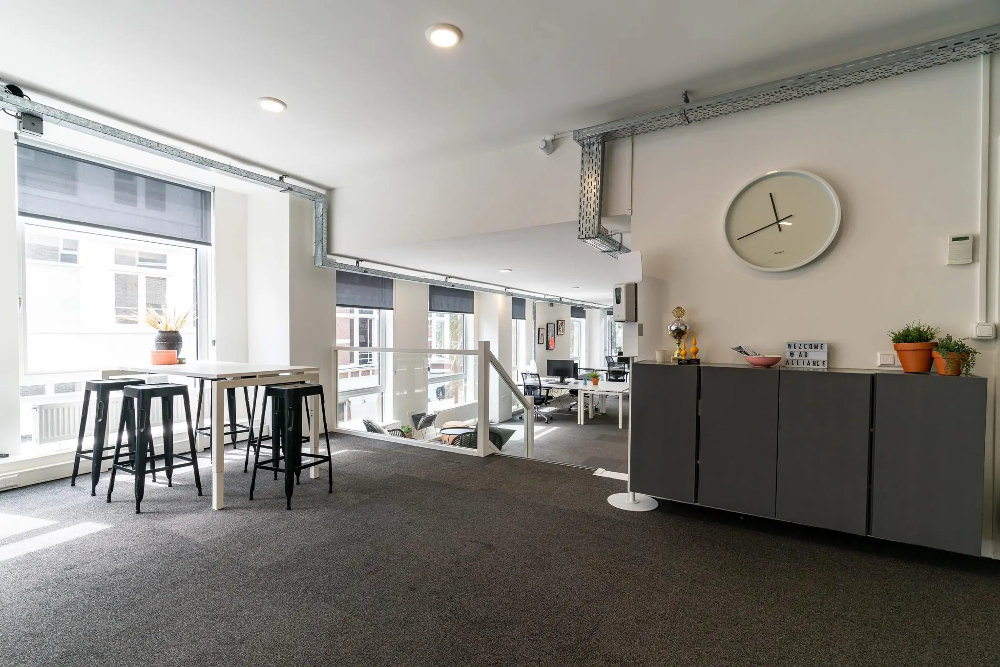 Moderne, open kantoorruimte met hoge ramen, een zithoek, planten en een wandklok.