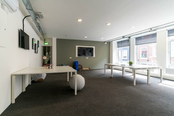 Moderne, lichte kantoorruimte met witte bureaus, grote ramen en een muurtelevisie.