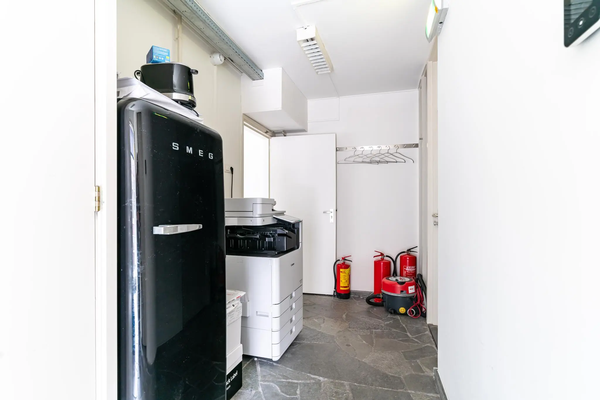 Kleine bedrijfsruimte met een zwarte SMEG-koelkast, een printer, brandblussers en schoonmaakapparatuur.