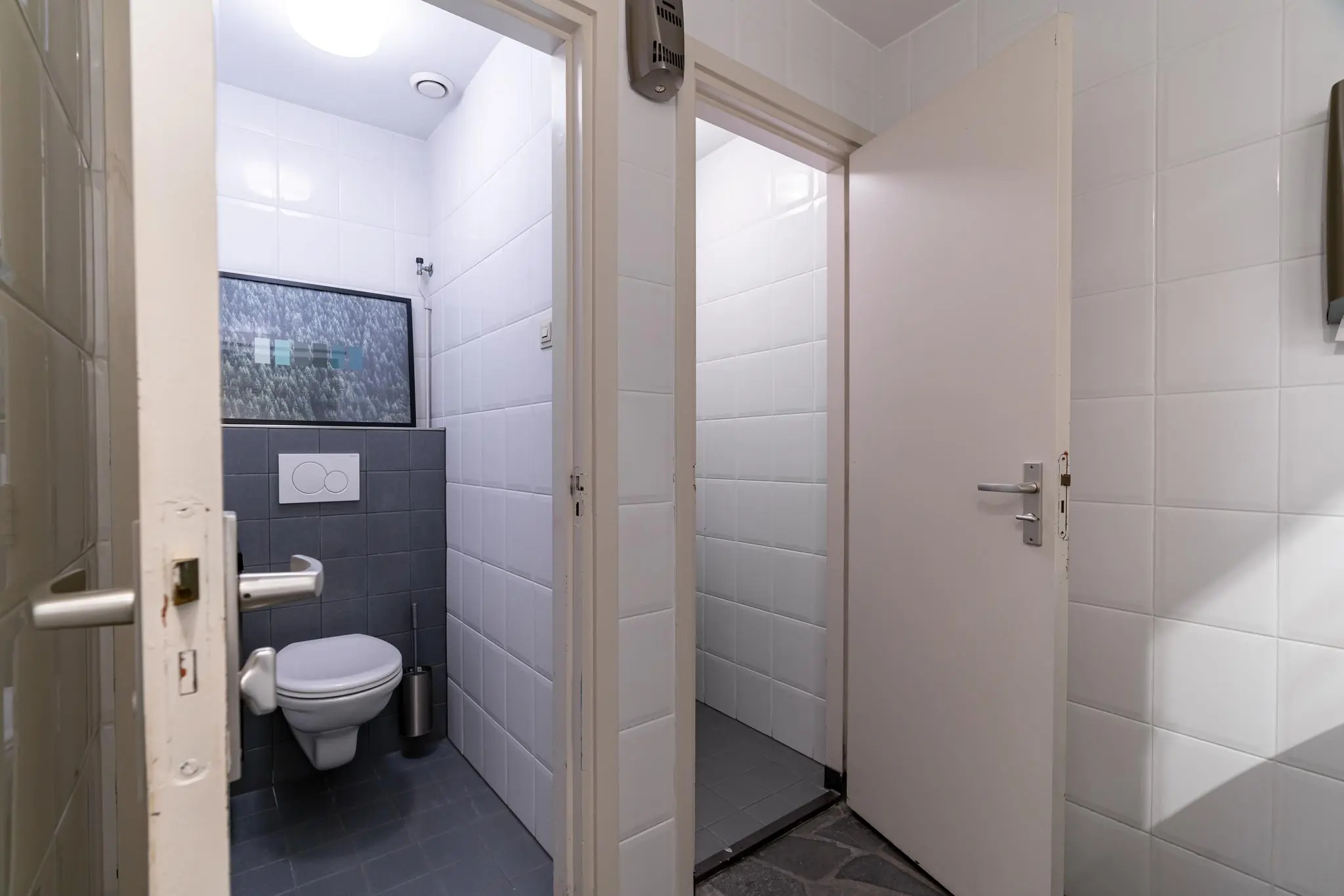 Twee toiletruimtes met witte tegels, waarvan één met een hangtoilet en kunstwerk aan de muur.