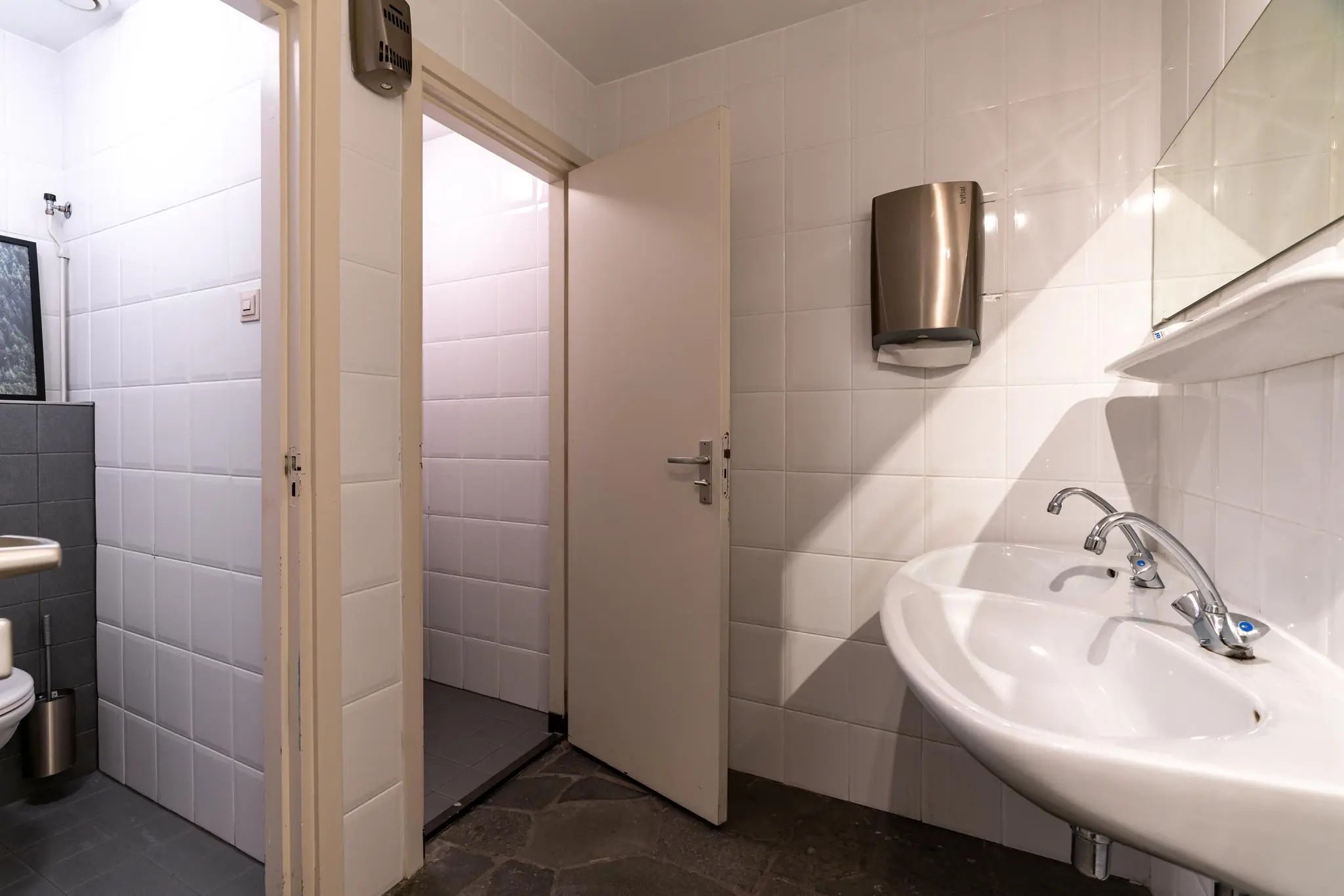 Wit betegelde openbare toiletruimte met twee wastafels, open deuren naar toiletcabines en een wandgemonteerde handdoekdispenser.