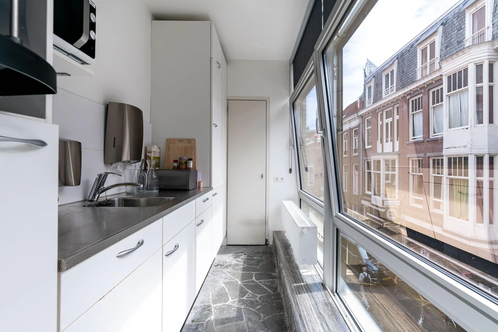 Smalle keuken met witte kasten en een groot raam met uitzicht op een straat en klassieke gevels.