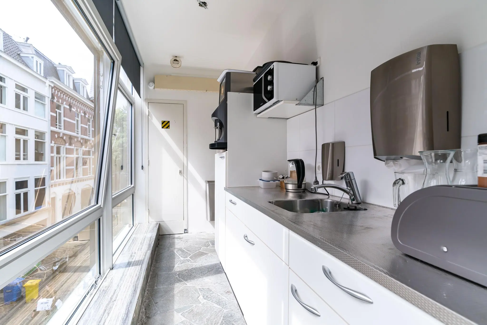 Moderne pantrykeuken met grote ramen, een koffiezetapparaat, magnetron en uitzicht op historische panden aan het Plein.