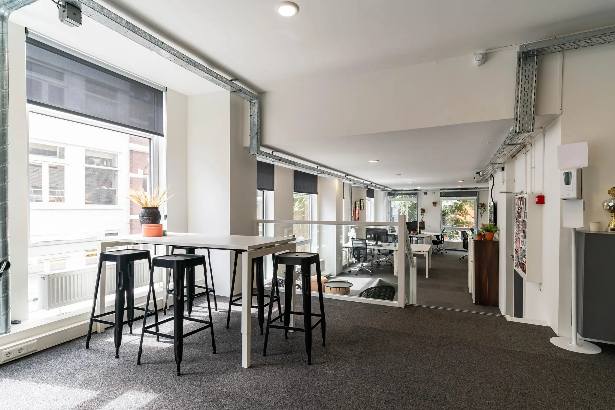 Moderne, lichte kantoorruimte met hoge tafels, barkrukken en werkplekken aan het raam.