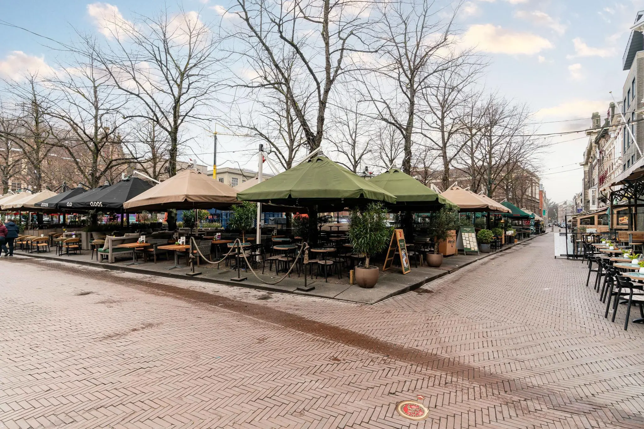 Een leeg terras met overkappingen op het Plein in Den Haag, omringd door kale bomen en restaurants.