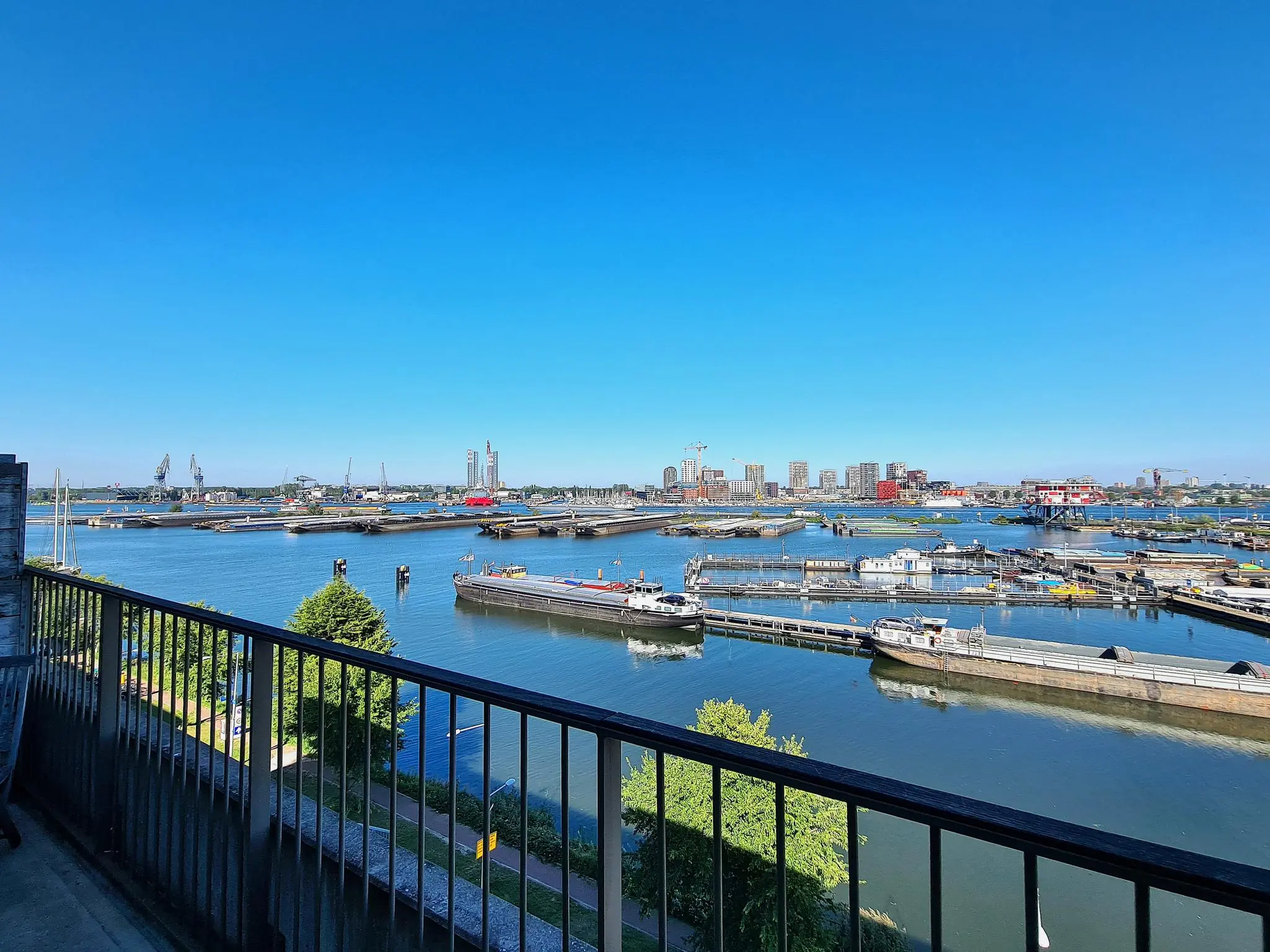 Uitzicht vanaf een balkon op de Danzigerkade over het IJ in Amsterdam, met schepen, kades en hoge gebouwen op de achtergrond.