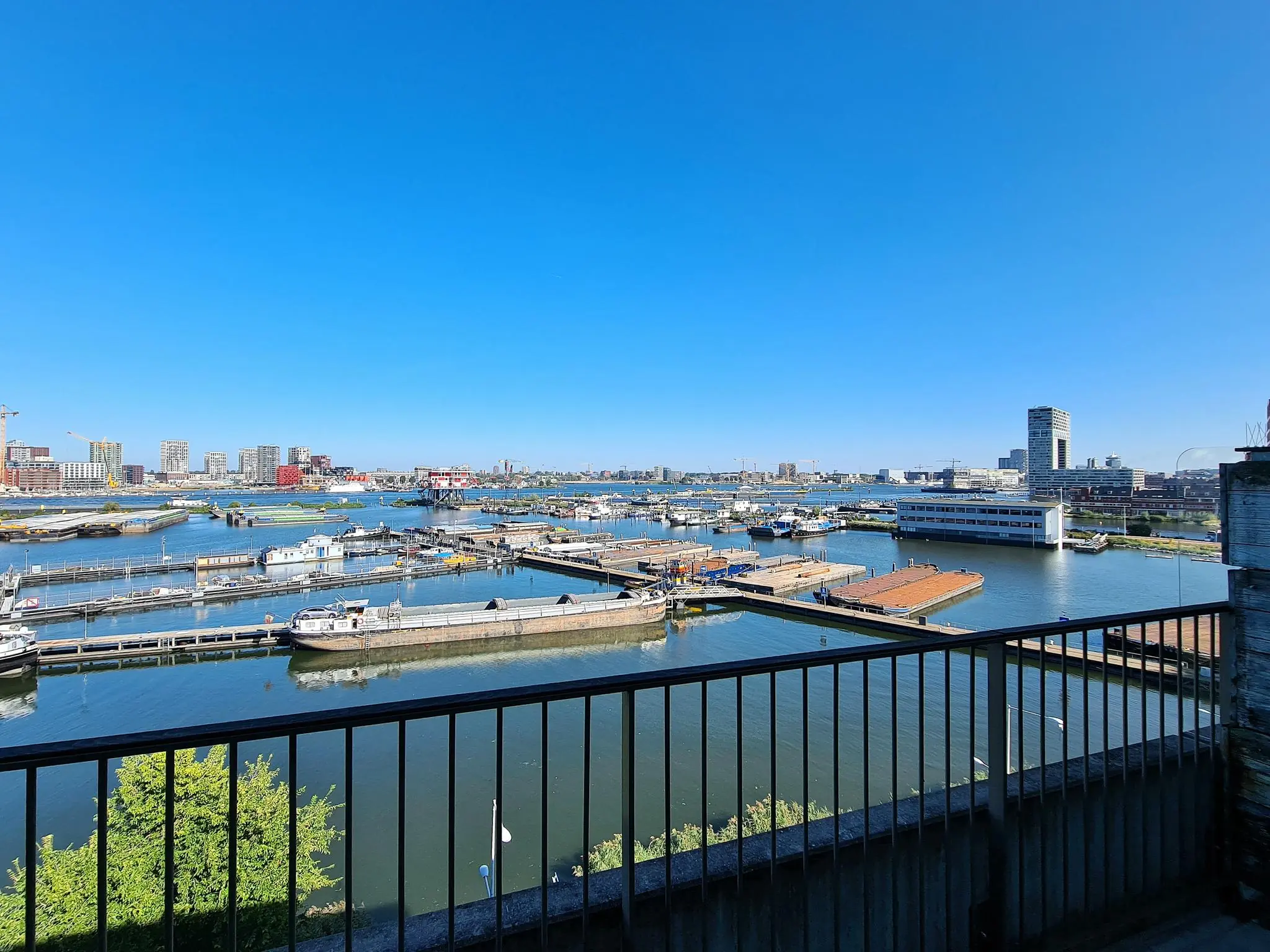 Uitzicht op de Danzigerkade in Amsterdam met een overzicht van de haven, boten en moderne gebouwen onder een heldere blauwe lucht.