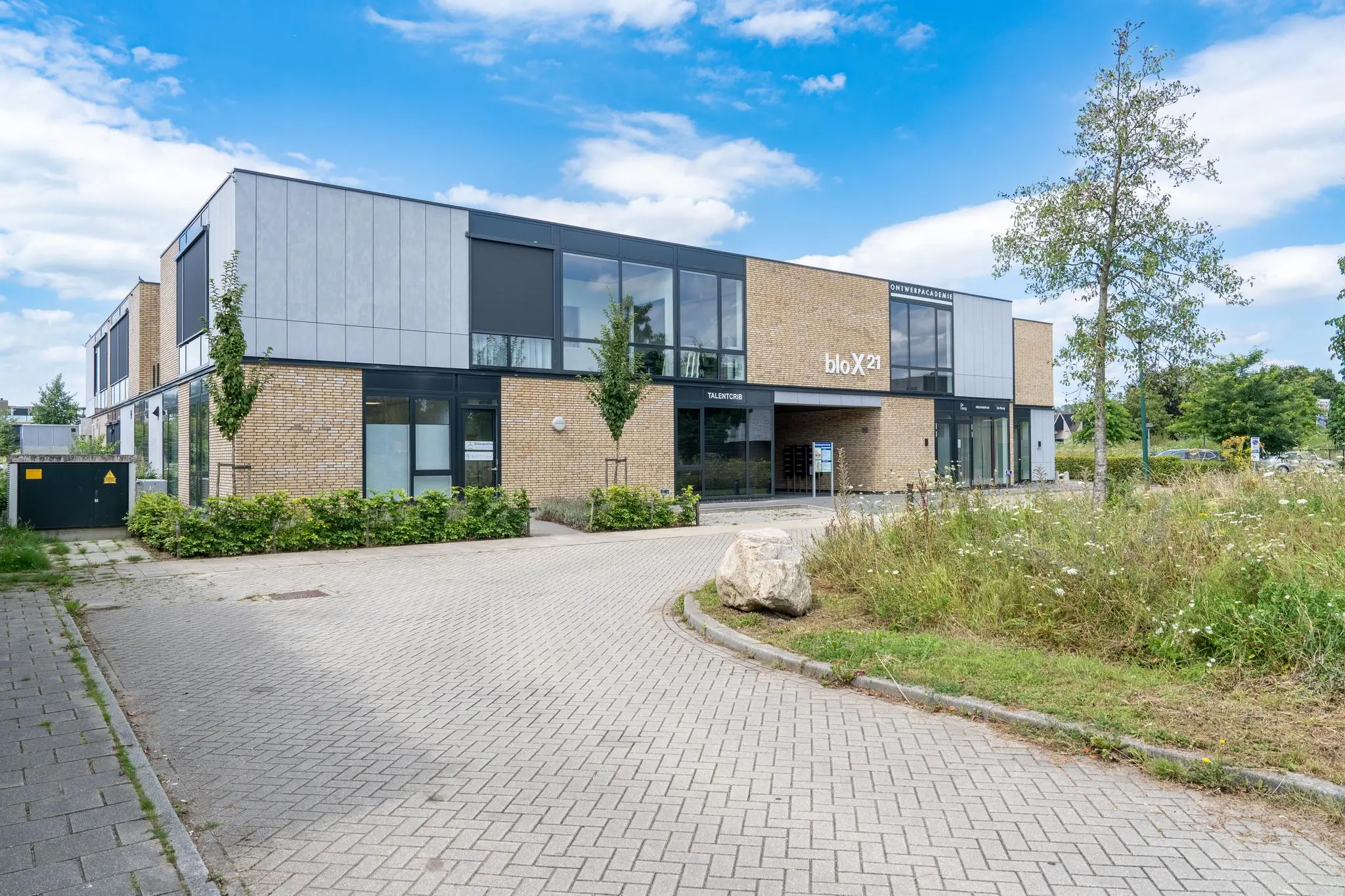Moderne bedrijfs- of onderwijsgebouw bloX21 met bakstenen gevel en grote ramen, gelegen aan een oprijlaan met groenstroken.