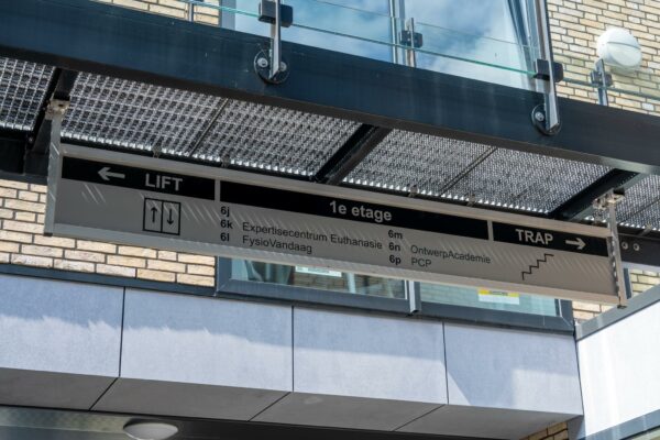 Wegwijzer op een gebouw met aanduidingen voor lift en trap naar de eerste etage met onder andere het Expertisecentrum Euthanasie en de OntwerpAcademie.