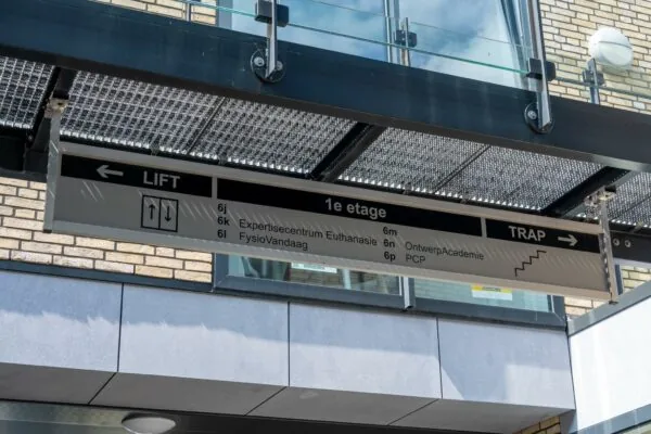 Wegwijzer op een gebouw met aanduidingen voor lift en trap naar de eerste etage met onder andere het Expertisecentrum Euthanasie en de OntwerpAcademie.