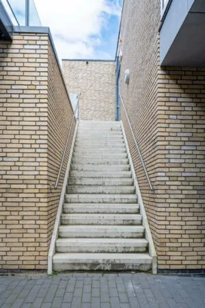Betonnen buitentrap tussen twee bakstenen muren met metalen leuningen, leidend naar een hoger gelegen niveau.