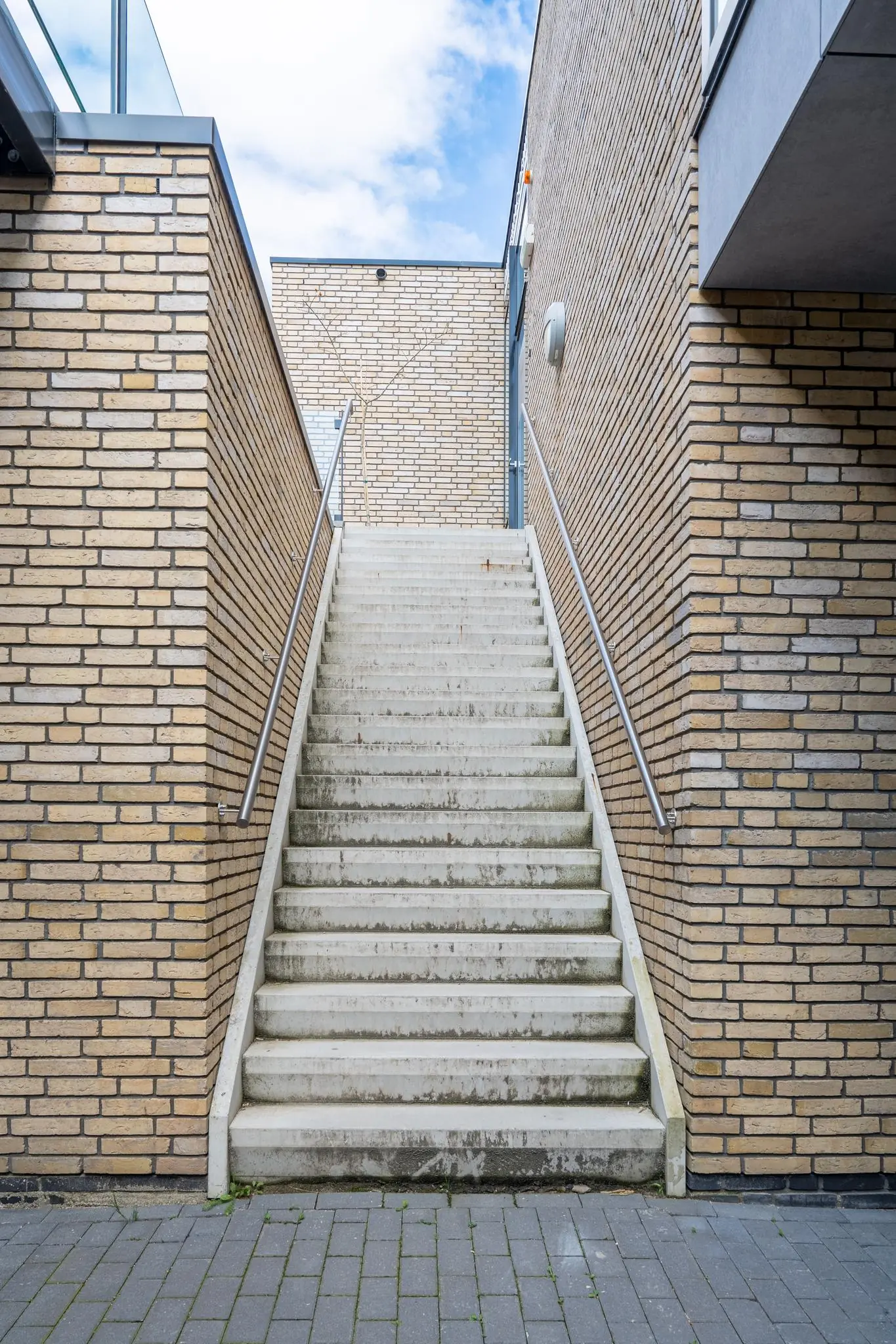 Betonnen buitentrap tussen twee bakstenen muren met metalen leuningen, leidend naar een hoger gelegen niveau.