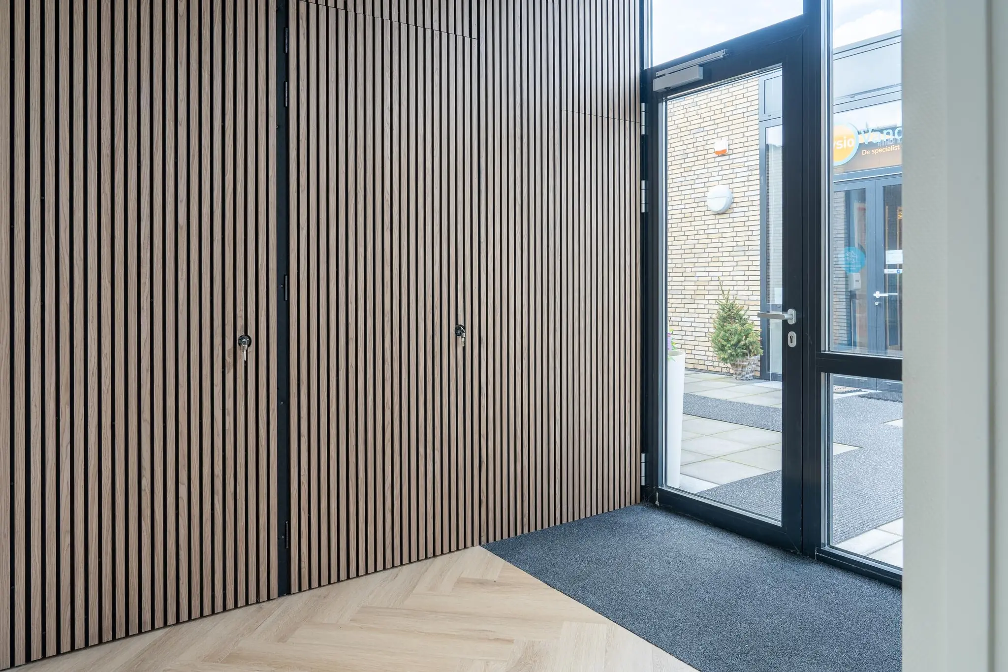 Ingang van een modern gebouw met houten lattenwand, glazen deur en zicht op een buitenruimte met bestrating.