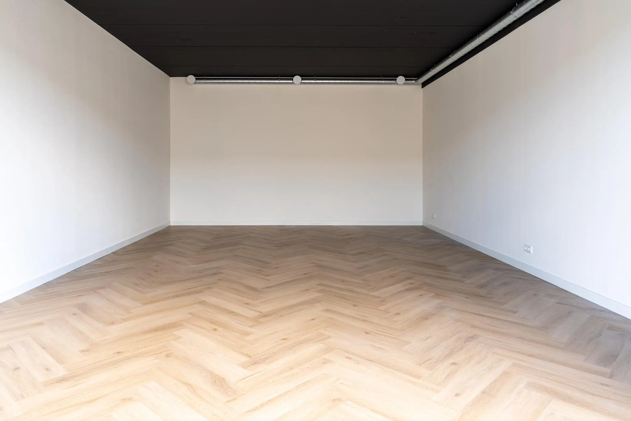 Lege kamer met visgraatvloer, witte muren en zwart plafond met zichtbare ventilatiebuis.