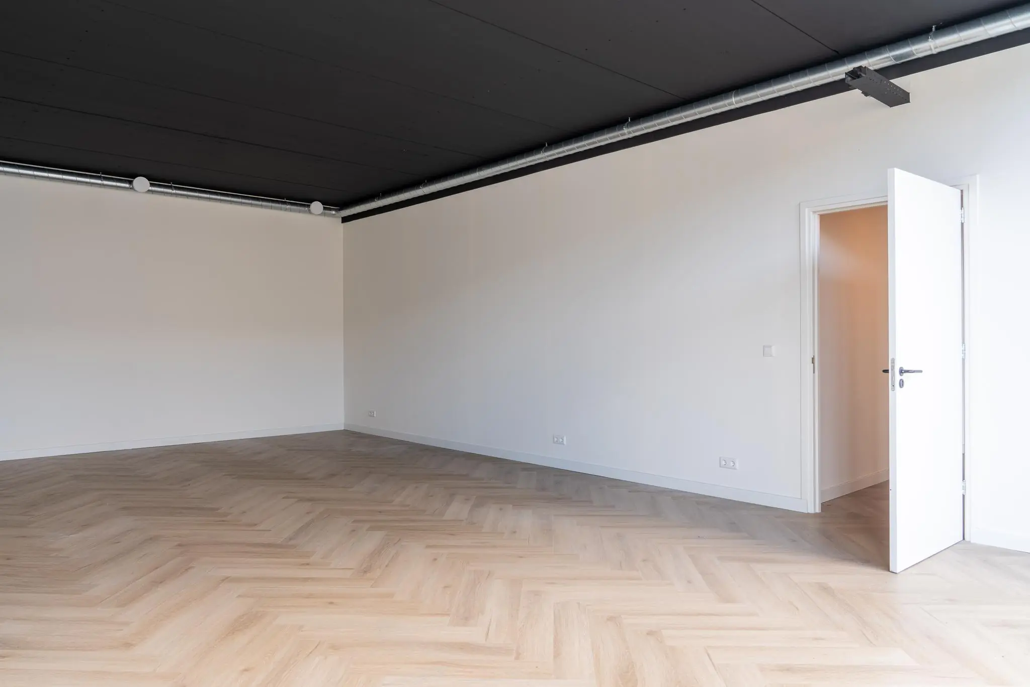 Lege kamer met visgraatvloer, witte muren, een zwart plafond met zichtbare ventilatiebuis en een openstaande witte deur.
