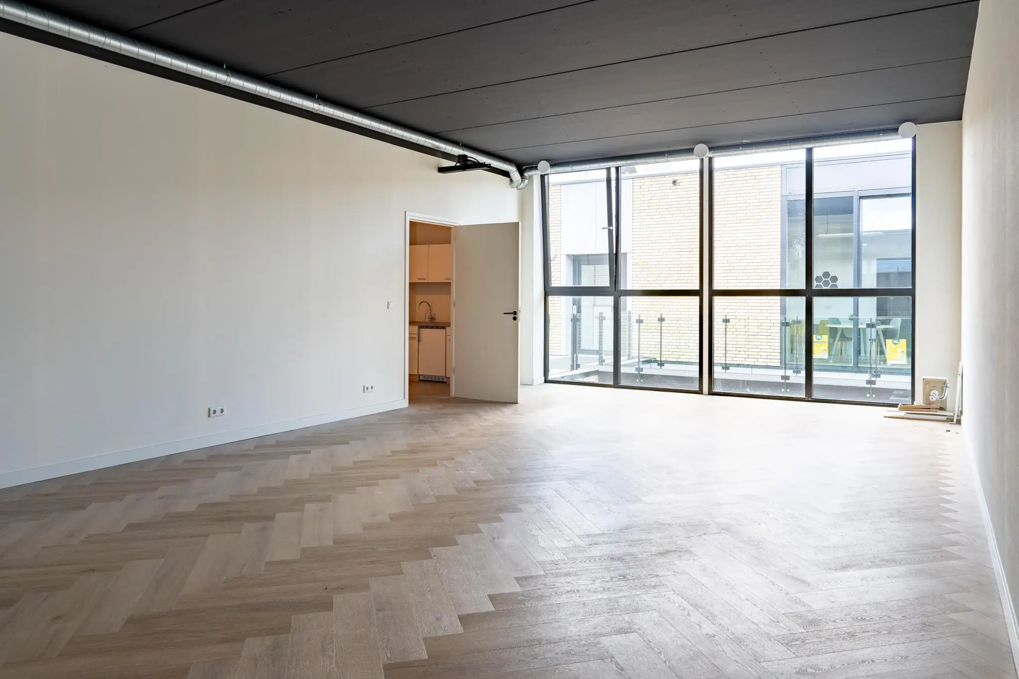 Lichte, lege woonkamer met visgraat vloer, zwarte plafonddetails en grote ramen met uitzicht op een binnenplaats.