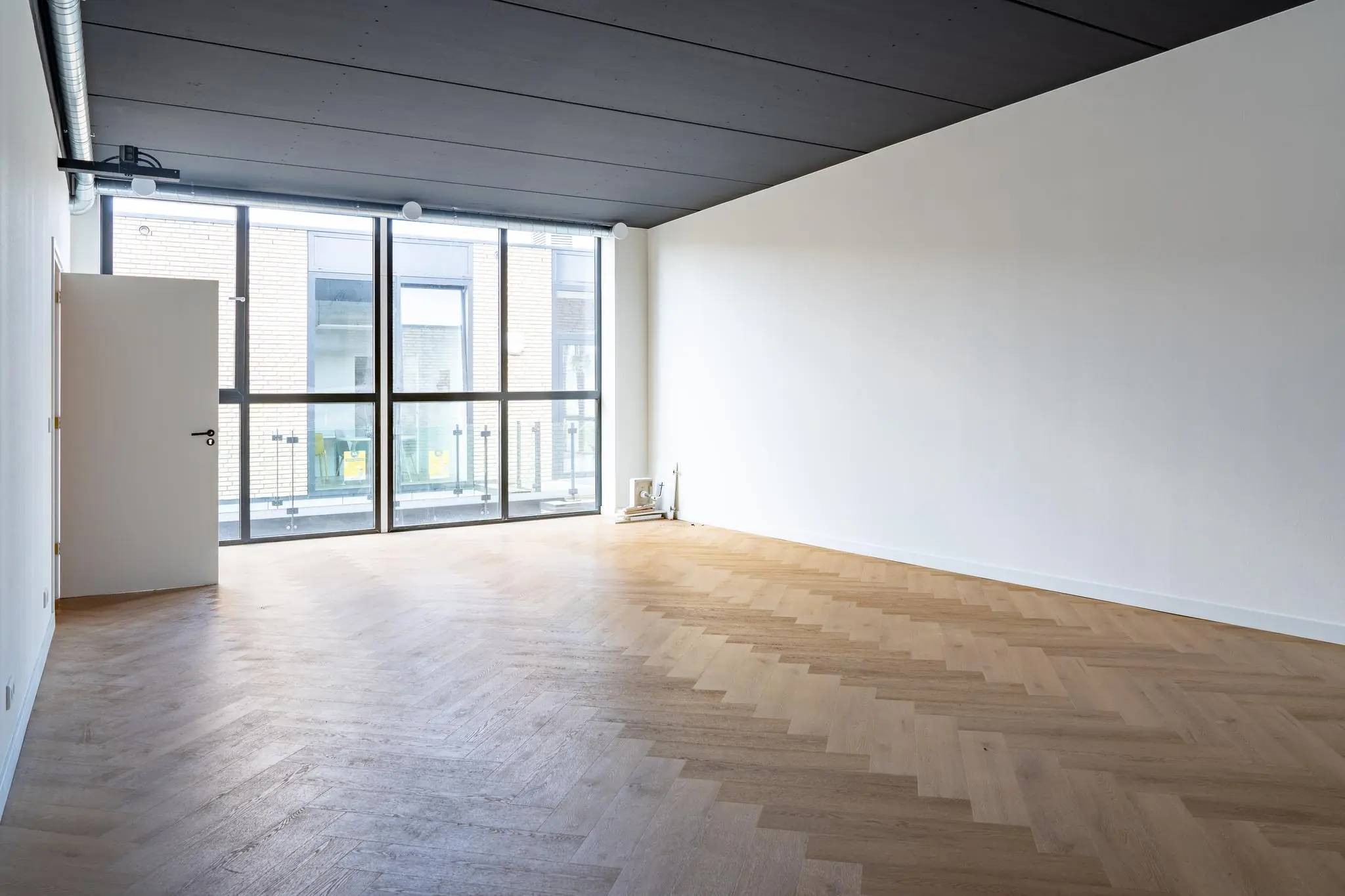 Lichte, lege kamer met visgraat houten vloer en grote ramen aan één zijde.