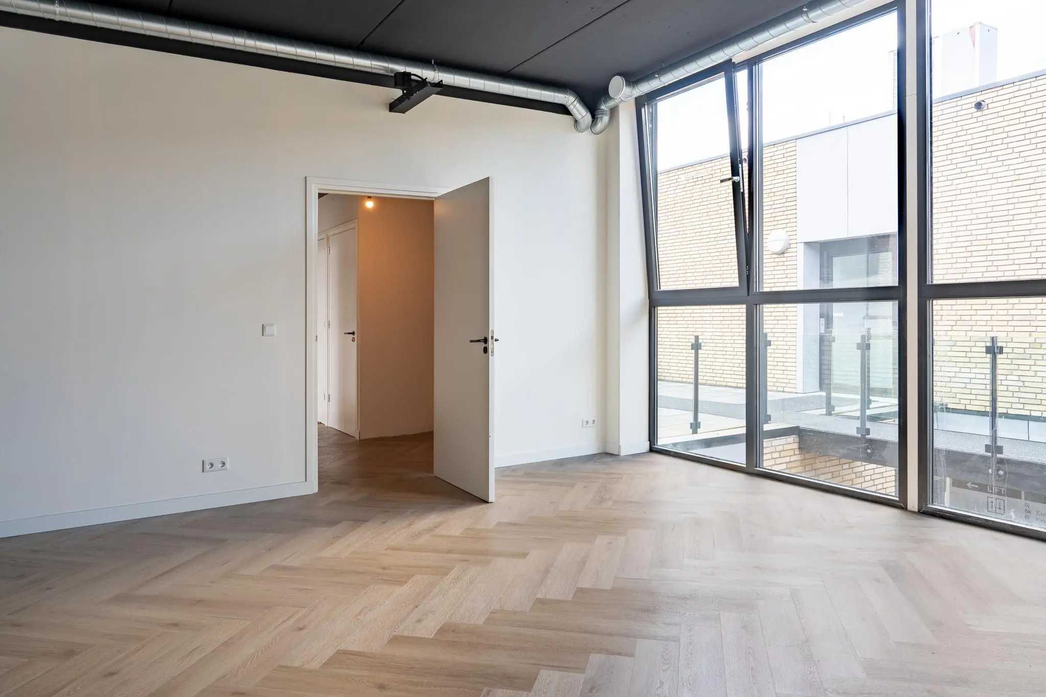 Lichte, lege kamer met houten visgraatvloer en grote ramen die uitkijken op een balkon met bakstenen gevel.